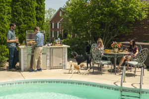 Outdoor Living Space: Friends sit on a patio with a pool, two men are grilling food.